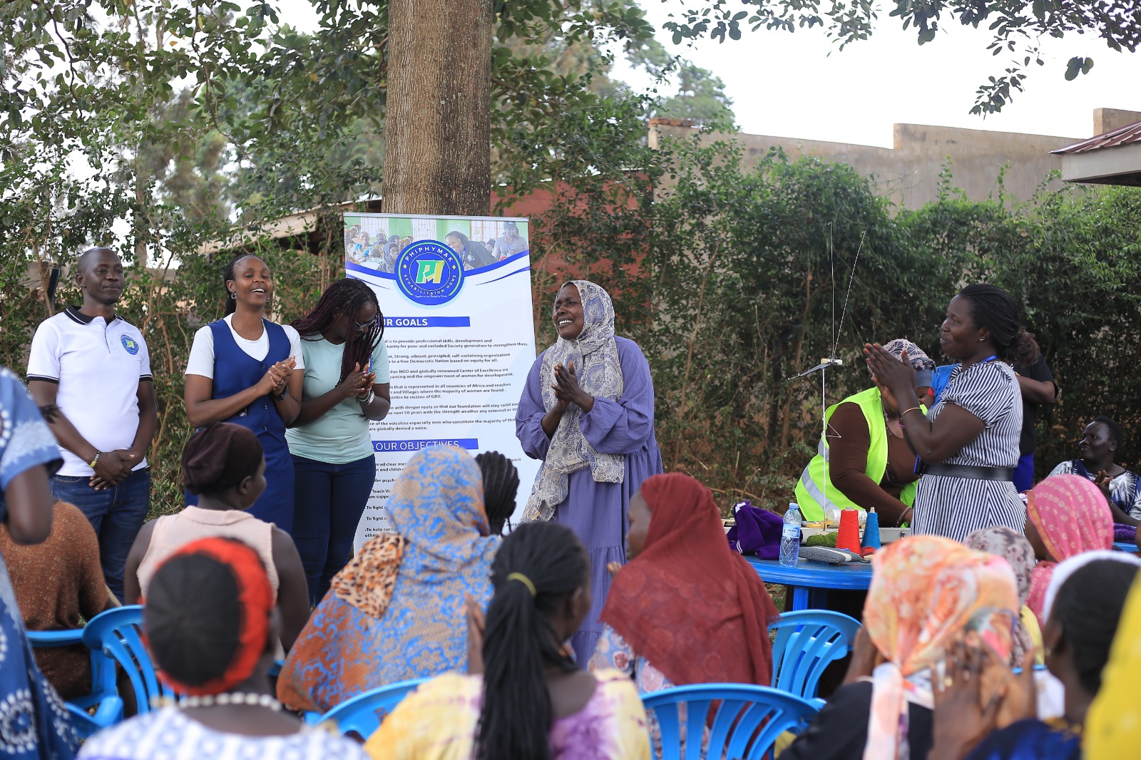 PhiphyMak team members and community volunteers standing beside the organisation banner as residents applaud during the outreach in Kiryandongo Refugee Settlement